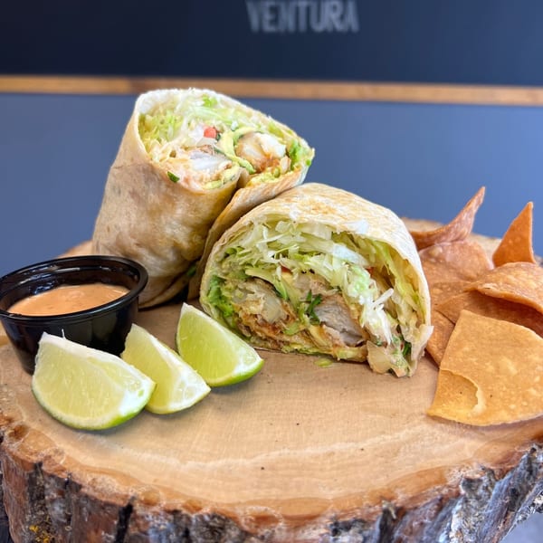 Two fish burritos, cut in half, with lime wedges, chipotle sauce, and tortilla chips on a wooden board.