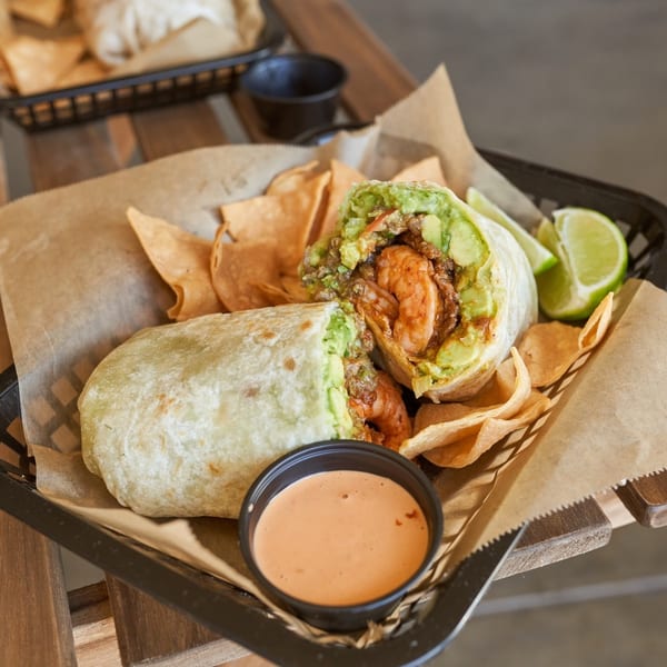 Two shrimp and avocado burritos with tortilla chips, lime wedges, and dipping sauce in a basket.