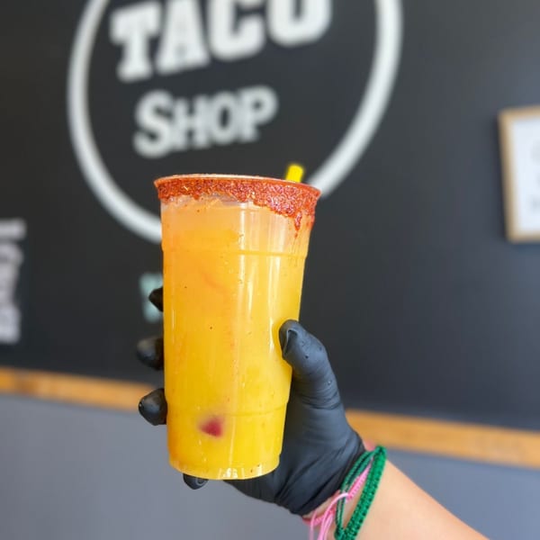 Hand in black glove holding a mango drink with chili rim against a blurred Taco Shop sign.