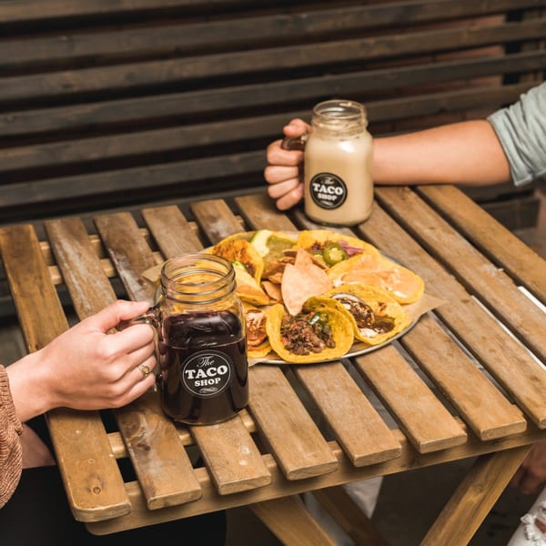 Hands holding branded mason jars with drinks, next to a platter of tacos and chips on a wooden table.