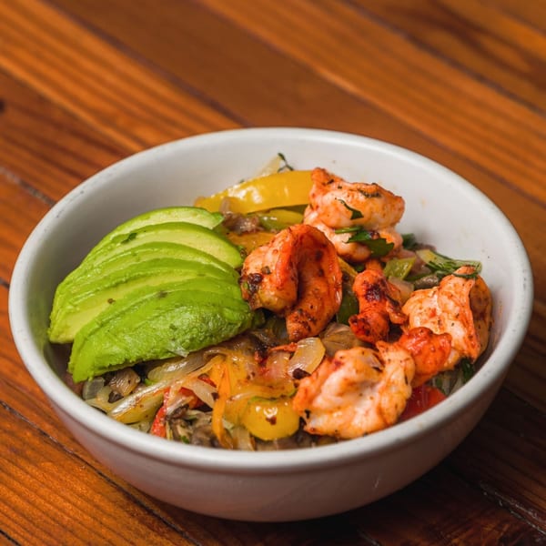 A white bowl filled with grilled shrimp, sliced avocado, and sautéed vegetables on a wooden table.