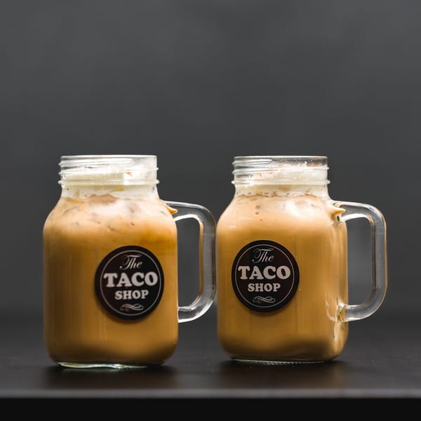 Two mason jar mugs filled with iced coffee and topped with foam, featuring "The Taco Shop" logo.