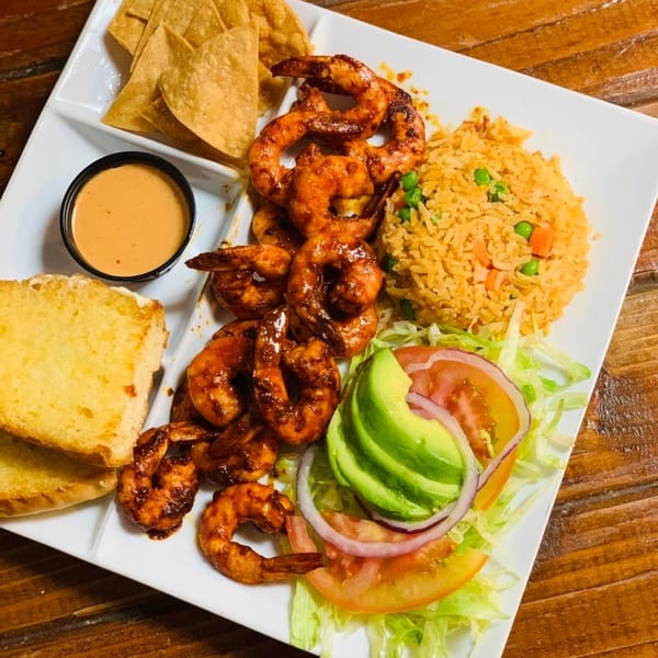 A Mexican shrimp dish with rice, salad, tortilla chips, and a side of sauce on a white plate.