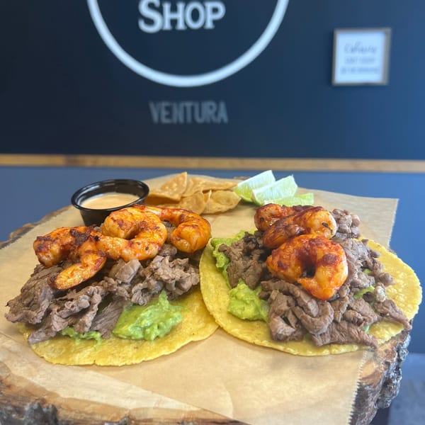Two carne asada and shrimp tacos with guacamole, chips, sauce, and lime wedges on a wooden board.