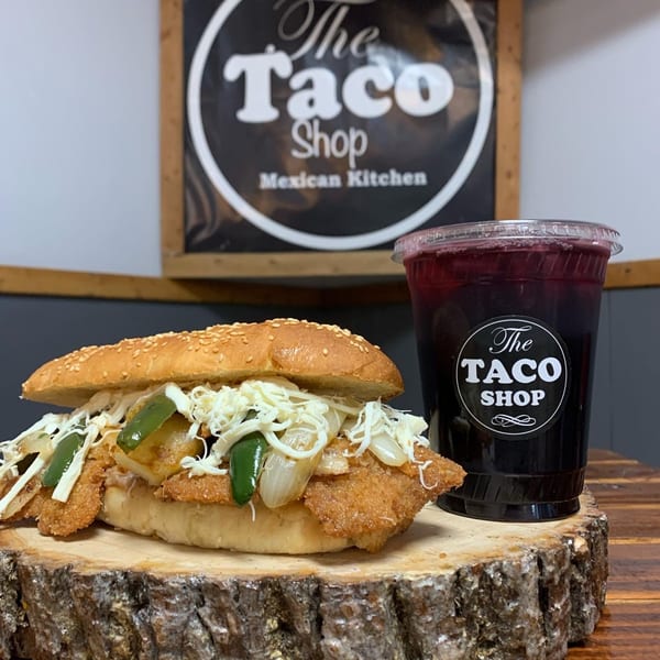 A Mexican torta with breaded chicken, cheese, onions, and peppers, next to a drink.