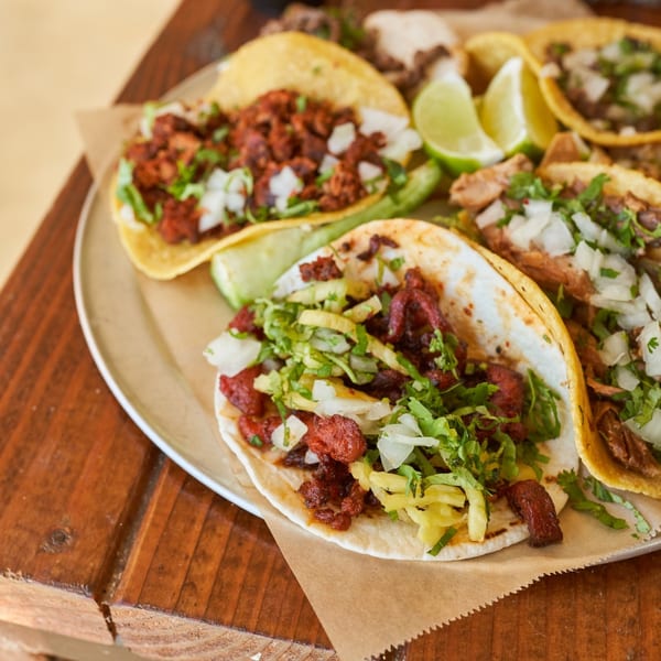 A metal tray of various Mexican tacos with meat, onions, cilantro, and lime wedges on a wooden table.